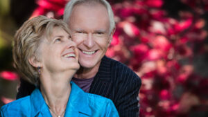 Two older people hold each other and smile in front of a red bush.