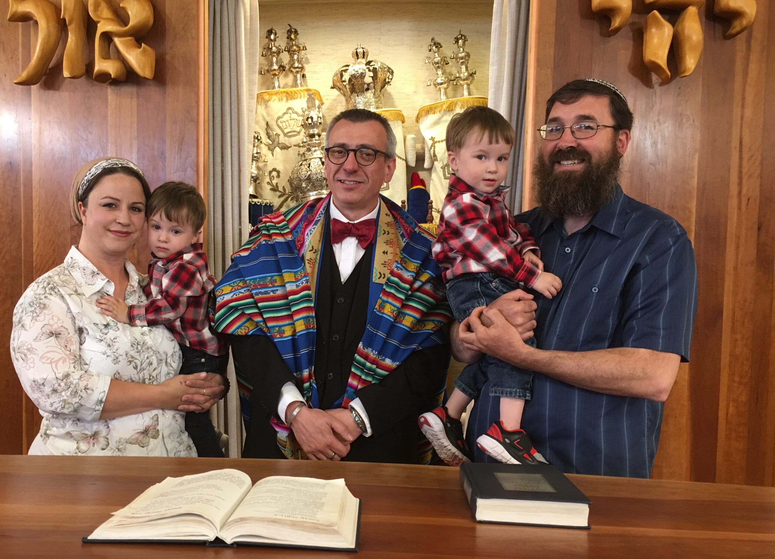 Two people hold their children while smiling, while a rabbi stands in the middle.