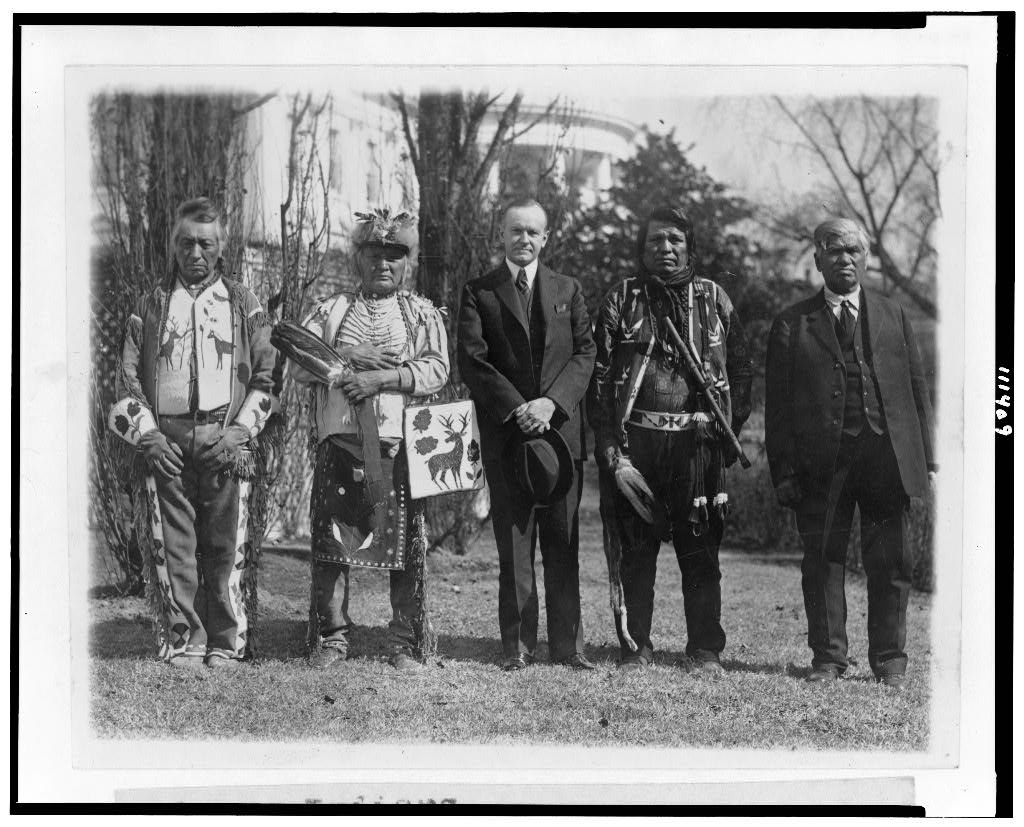 TheVoteEp3Option2 A man in a suit is flanked by Indigenous representatives in assorted regalia, including one in a suit.
