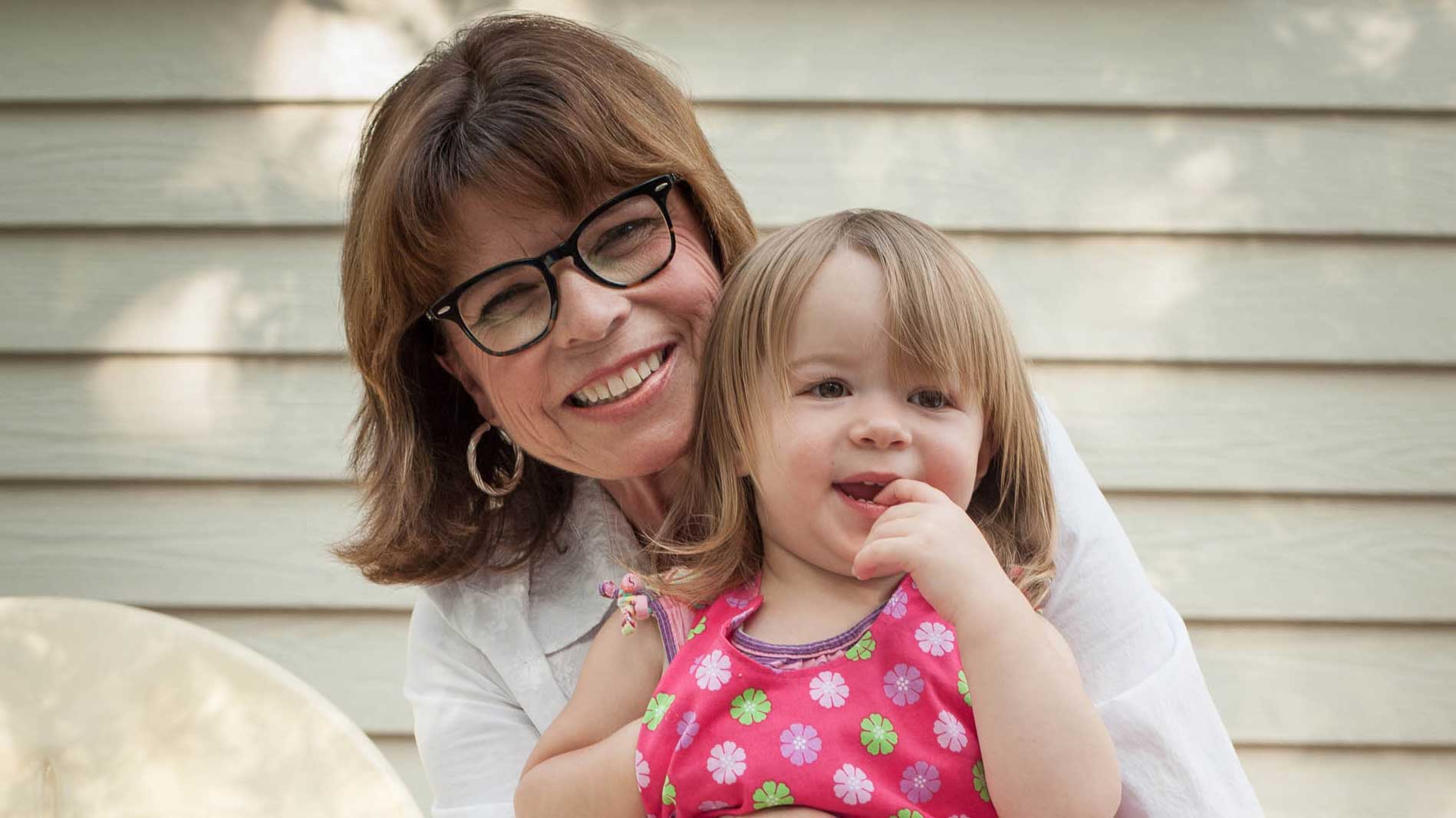 A woman with glasses smiles as she holds a toddler, who is also smiling.